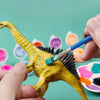 A hand holds a yellow dinosaur model being painted with various colored paints.