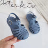 A hand holds a pair of blue Gladiator jelly sandals with woven straps and a non-slip sole.