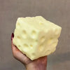 A pale yellow cheese wedge squishy stress relief toy is held in a hand against a neutral background.