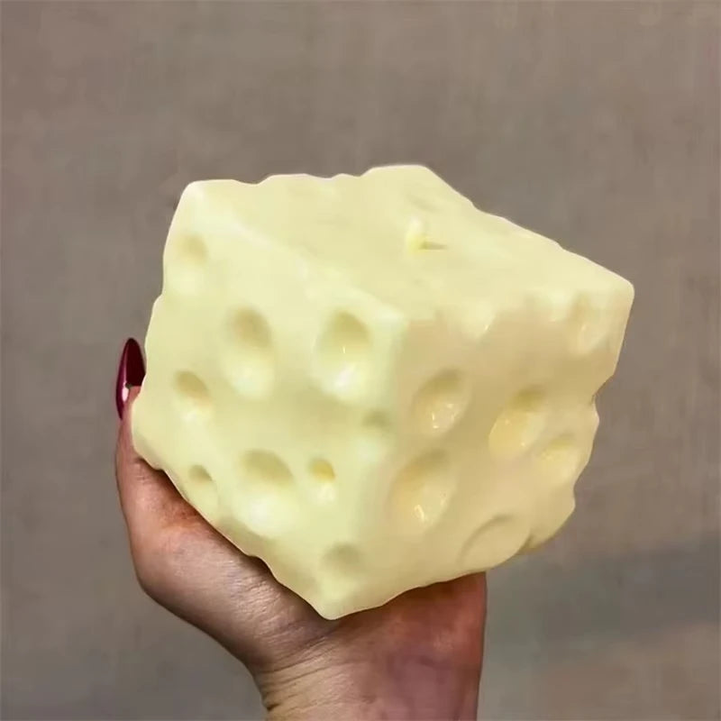 A pale yellow cheese wedge squishy stress relief toy is held in a hand against a neutral background.