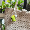 A handmade crochet dinosaur keychain hanging from a beige woven fabric bag.