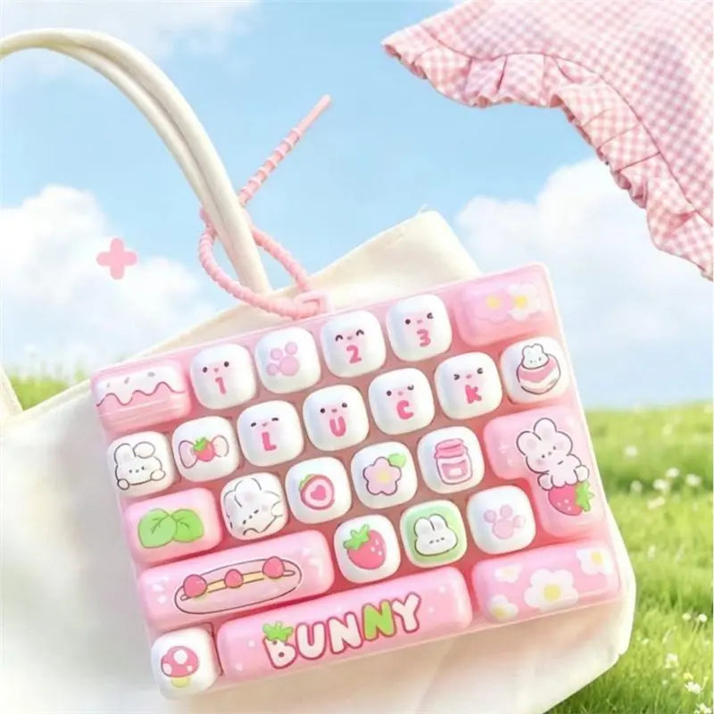 A pink and white cartoon animal keyboard keycap set is displayed on a white bag in an outdoor setting.