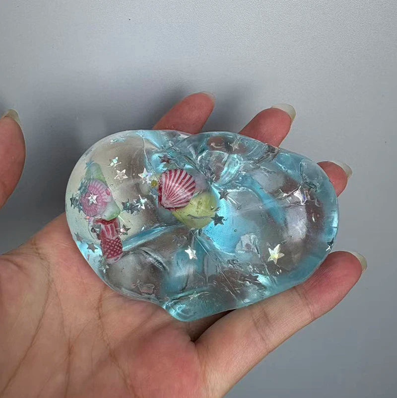 A hand holds a clear blue glass heart-shaped block squishy stress ball decorated with stars and seashells.