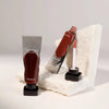 A bottle of Dark Mirror Lip Glaze is displayed next to a white rock, showing the product packaging and applicator.