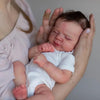A newborn baby is being held by a person, showing the realistic silicone doll with hand-rooted hair.