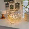 A clear glass cube night light filled with glowing white tulip flowers sits on a white surface.