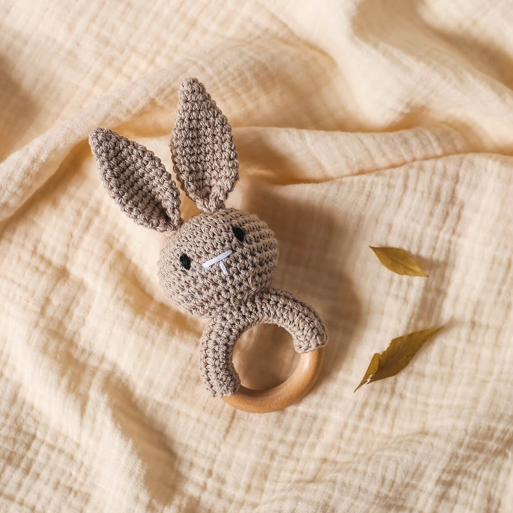 A handmade crochet bunny rattle set with soft gray ears and a wooden ring, resting on a light beige fabric.