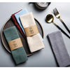 A set of classic cotton-linen dinner napkins in various muted colors, including beige, blue, green, and gray, arranged on a w