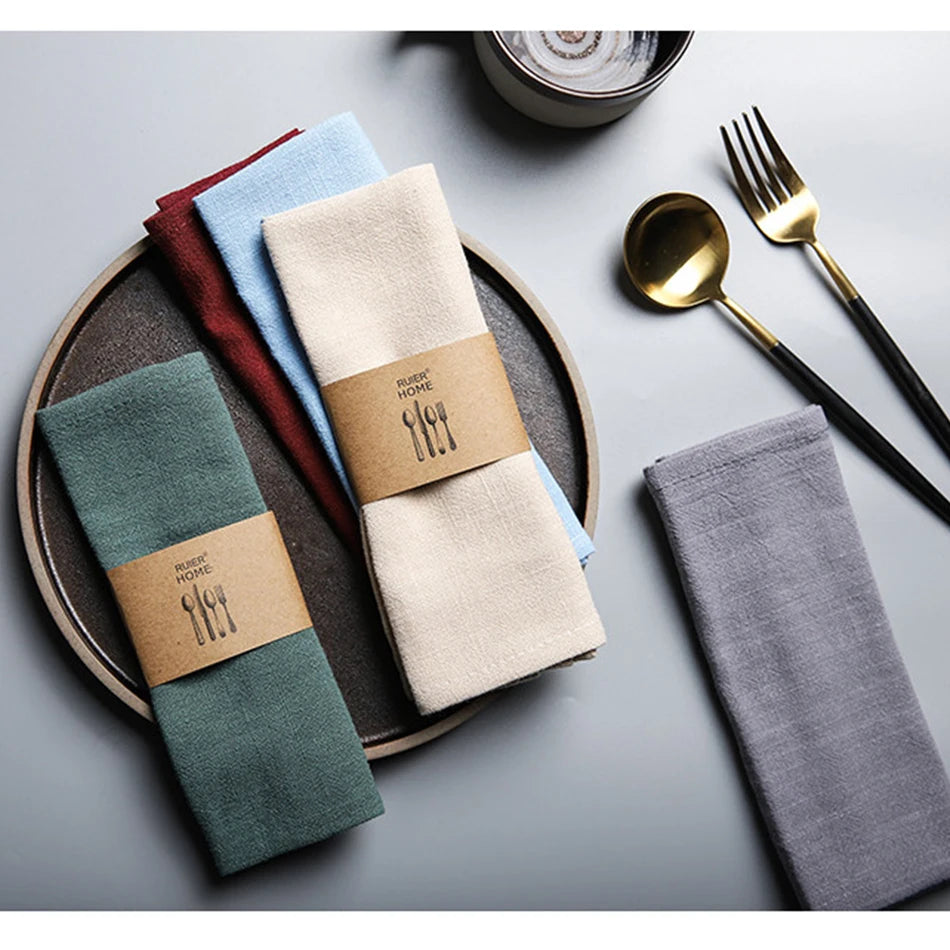 A set of classic cotton-linen dinner napkins in various muted colors, including beige, blue, green, and gray, arranged on a w