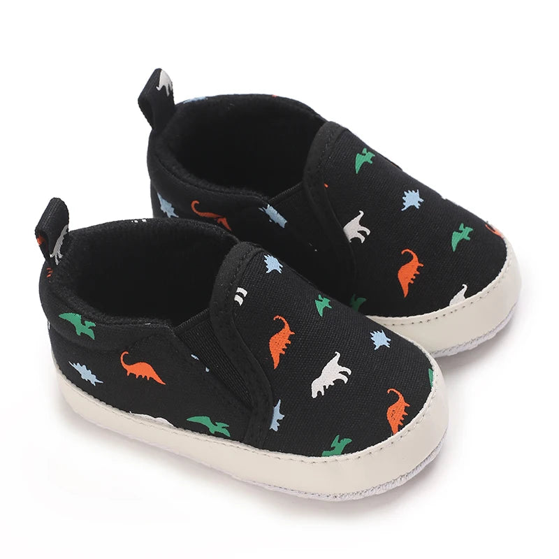 A pair of black baby first walker sneakers featuring a cute dinosaur print with green and white accents.