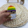 A hand holds a crocheted potato ornament with colorful yarn hair and a tag on a woven mat.
