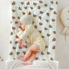A baby wearing a beige fleece beanie and yellow outfit is lying on a white blanket patterned with brown bear shapes.