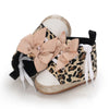 A pair of leopard print baby sneakers with pink bow accents and gold glitter soles.