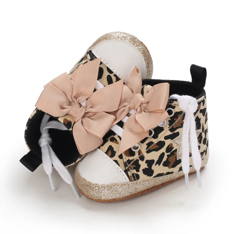 A pair of leopard print baby sneakers with pink bow accents and gold glitter soles.