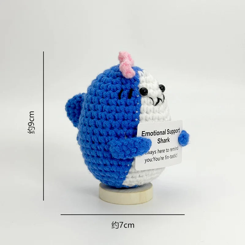 A handwoven crochet ornament depicting a blue shark with a pink fin and a white shark, used as desk decor.
