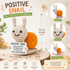 A handmade crochet positive snail toy in light gray and orange with a motivational card.