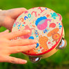A hand holds a round wooden tambourine with a colorful cartoon design featuring hearts and various animals.