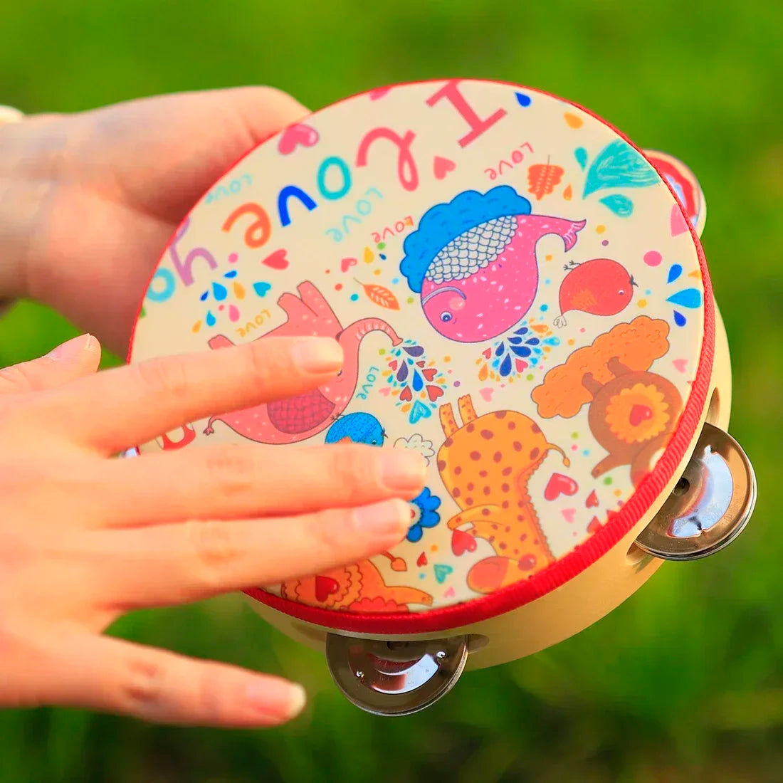 A hand holds a round wooden tambourine with a colorful cartoon design featuring hearts and various animals.