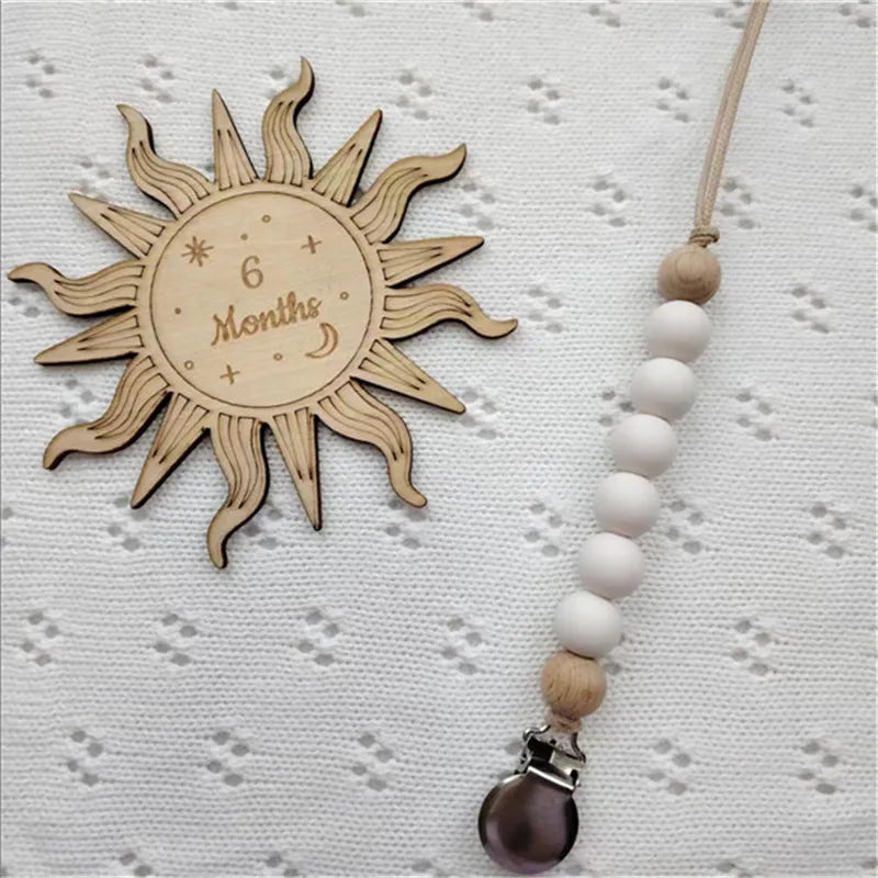 A wooden sun-shaped bead pacifier clip with six months marking and a silicone teether on a light fabric.