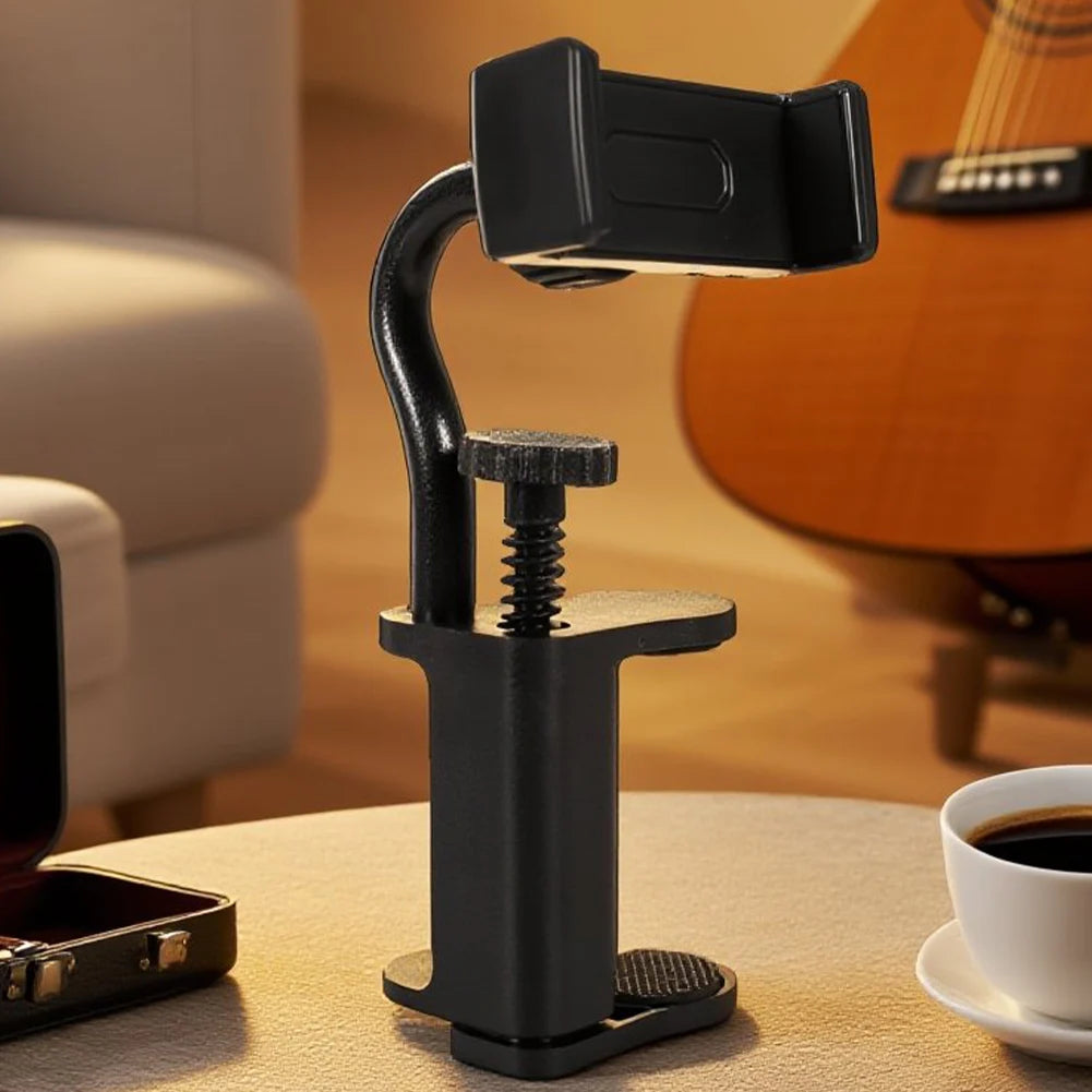 A black hands-free guitar phone holder with an adjustable clamp resting on a wooden table.