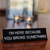 A black desk sign with white text that reads I'M HERE BECAUSE YOU BROKE SOMETHING