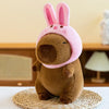 A brown capybara plush toy wearing a pink hood, sitting on a woven mat.