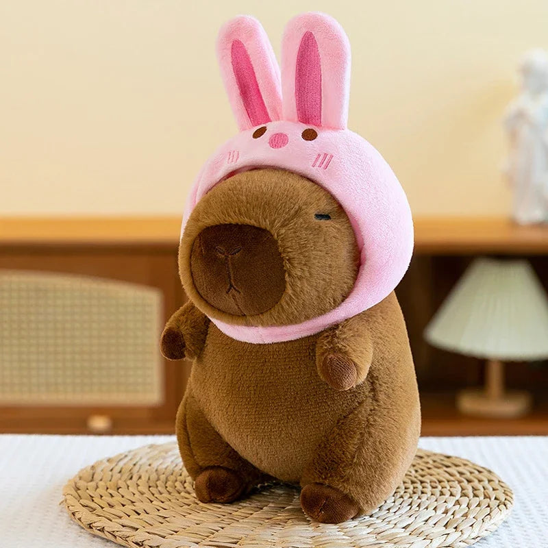 A brown capybara plush toy wearing a pink hood, sitting on a woven mat.