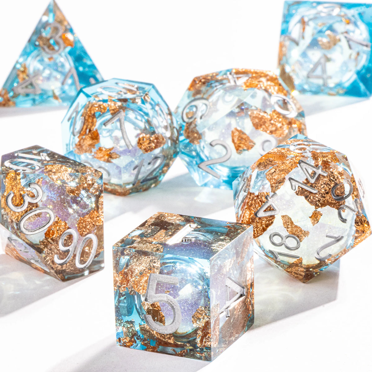 A collection of polyhedral dice in shades of blue and gold with a galaxy finish scattered on a white surface.