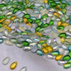 A close-up view of various translucent green, yellow, and clear water drop Czech glass beads scattered on a white surface.