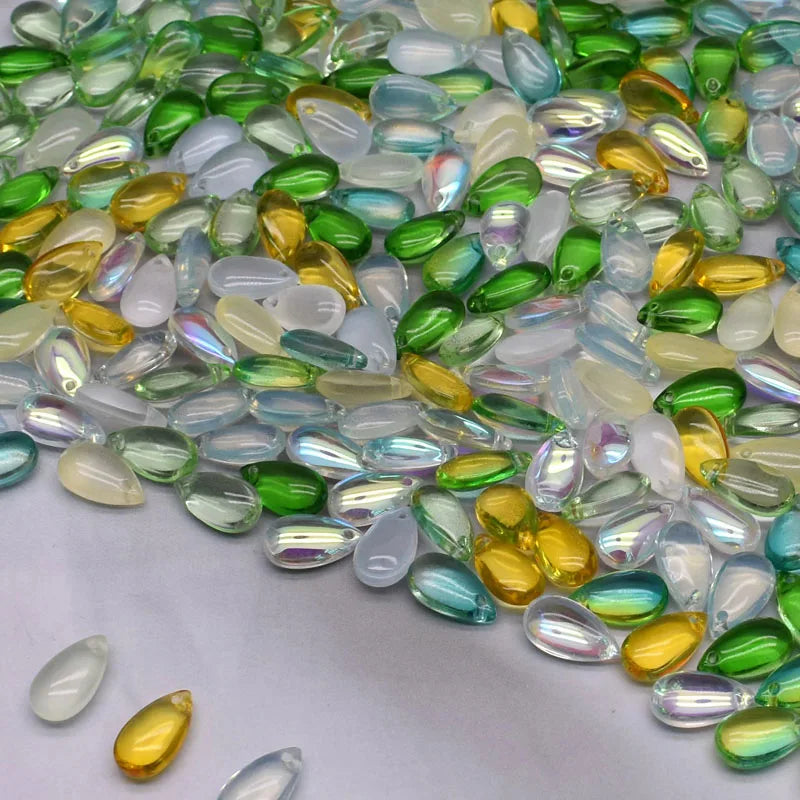 A close-up view of various translucent green, yellow, and clear water drop Czech glass beads scattered on a white surface.