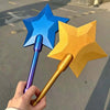 A hand holds a blue and yellow star-shaped magic wand with a purple handle against an asphalt background.