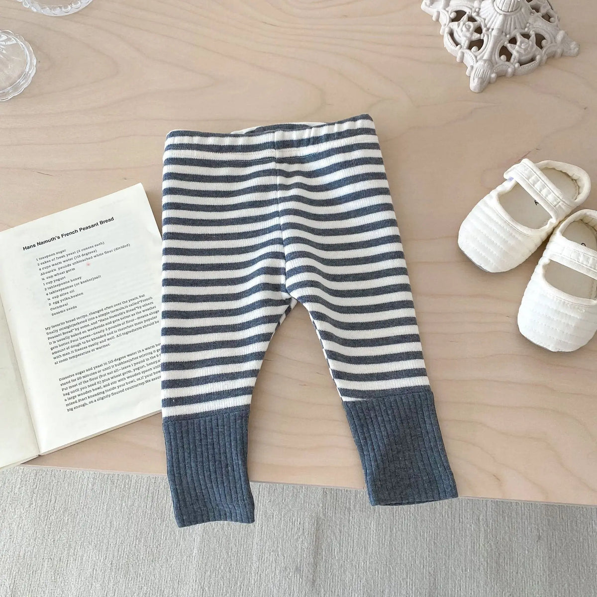 A pair of navy and white striped baby leggings lying on a wooden surface next to a book and white shoes.