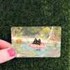 A card featuring a cute black cat floating in water surrounded by colorful flowers and foliage.