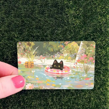 A card featuring a cute black cat floating in water surrounded by colorful flowers and foliage.