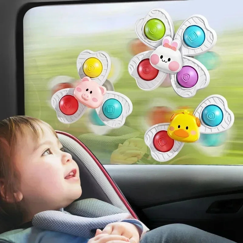A baby is looking at a window showing three colorful flower-shaped bath toys with suction cups and spinners.