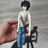 A hand holds a poseable L Lawliet plush doll from Death Note, featuring dark hair and blue clothing.