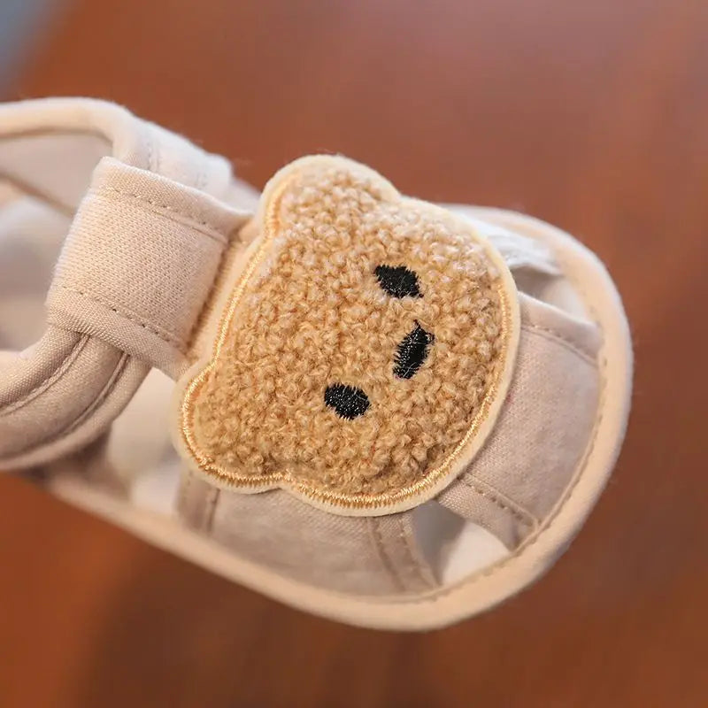 A pair of beige soft sole first walker shoes for babies featuring a cute bear face patch.