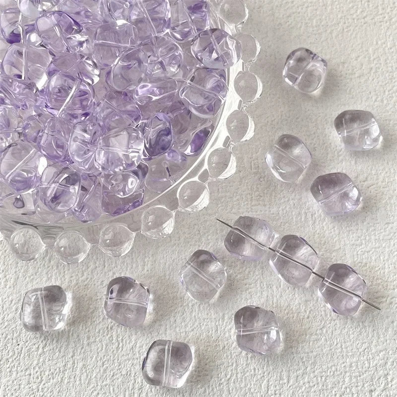 A pile of irregular square geometric glass beads in light purple are scattered on a white surface.