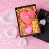 A set of pink silicone heart candle molds, including a large heart and a smaller heart, displayed in a black box on pink conf