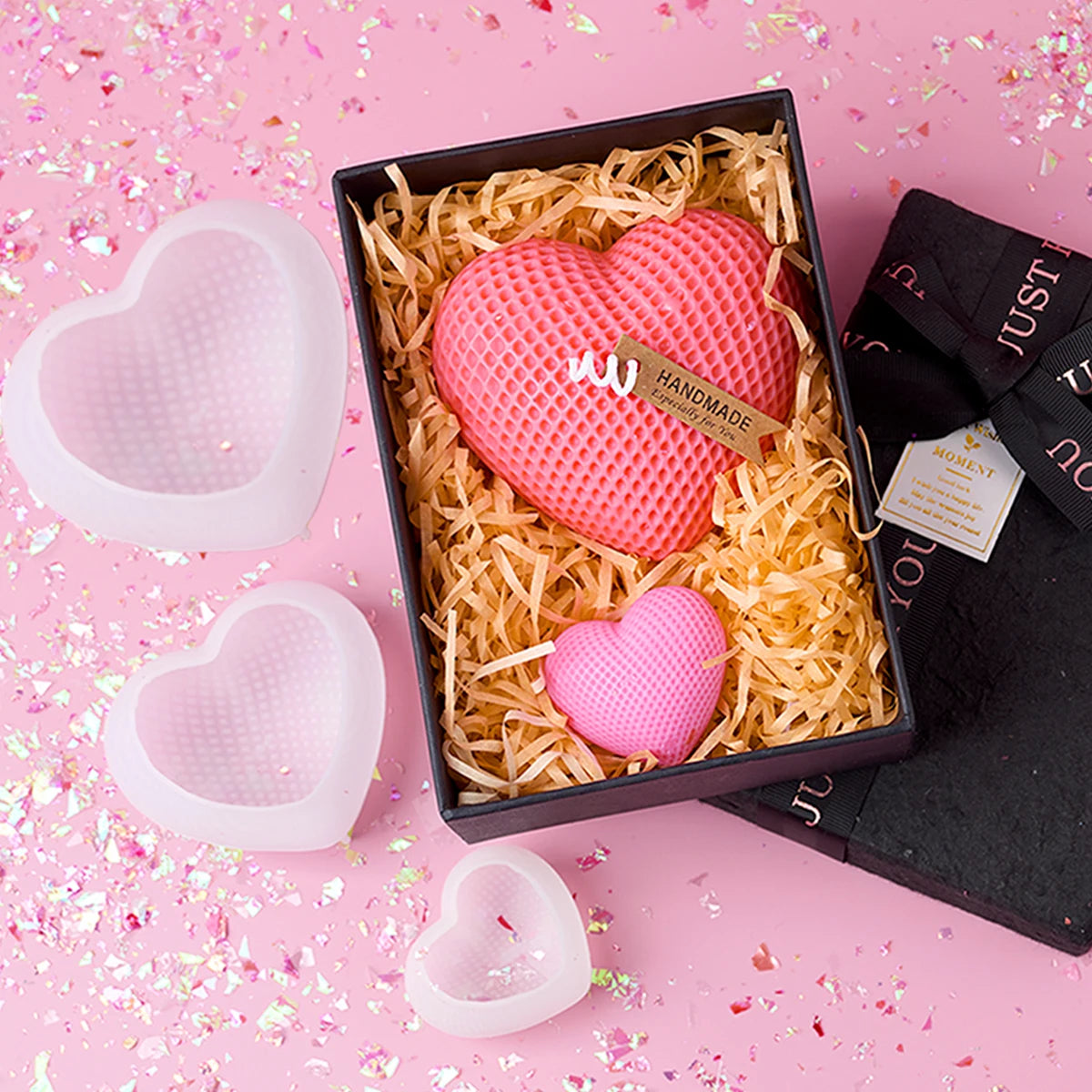 A set of pink silicone heart candle molds, including a large heart and a smaller heart, displayed in a black box on pink conf