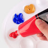 A hand is applying red acrylic paint to a white palette, with separate blobs of yellow and blue paint visible.