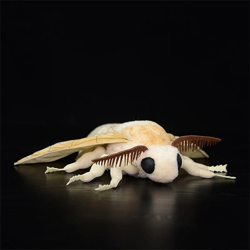 A soft, white and orange silk moth plushie with brown wings is lying on a dark surface.