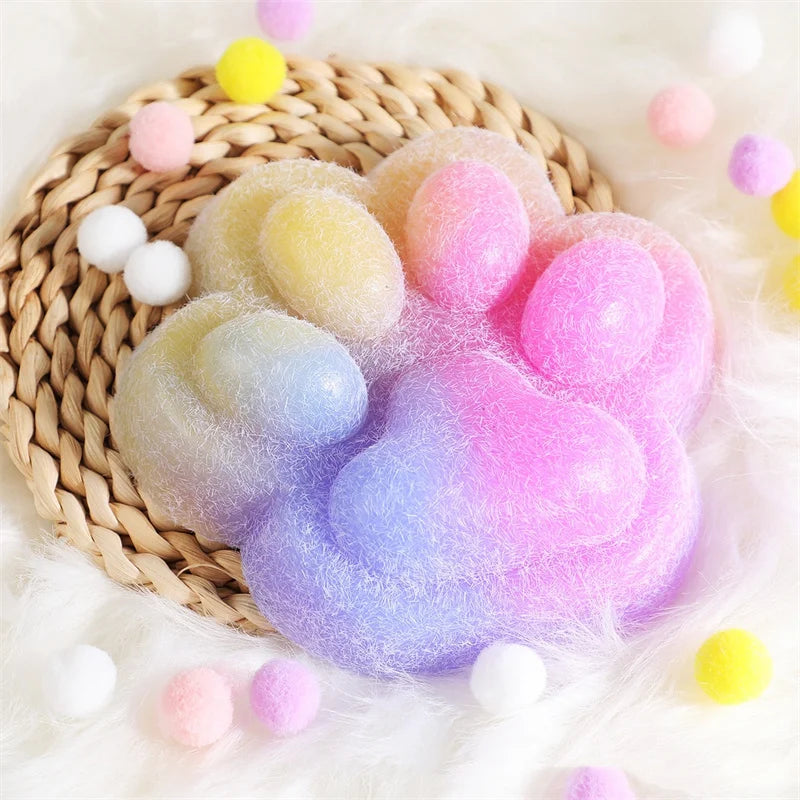 A giant pink and purple squishy cat paw toy resting on a woven mat surrounded by fluffy white and colorful pom-poms.