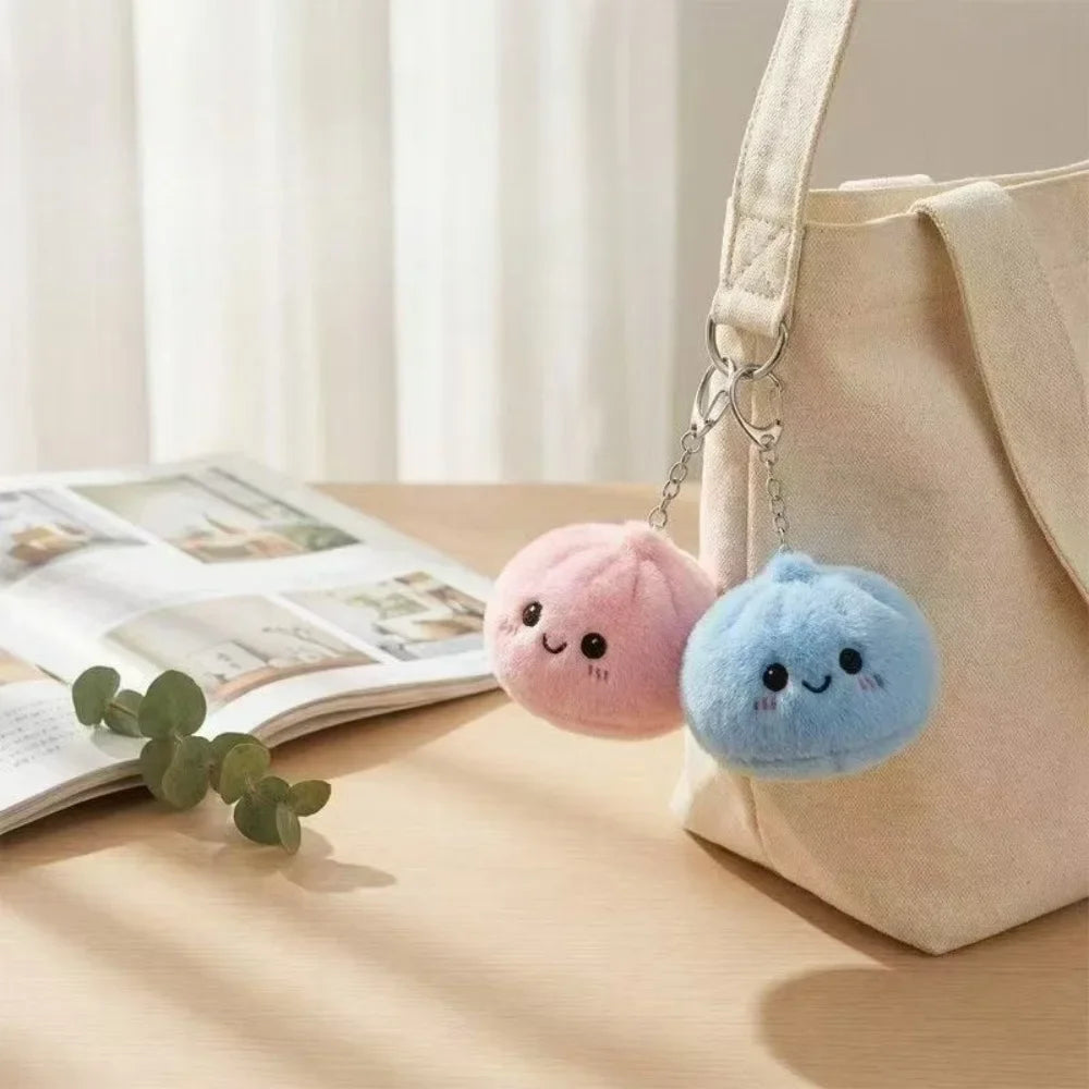 A light beige fabric pouch with a strap holds two round, squishy plush keychains in pink and blue.