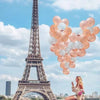 A cluster of pink and white balloons is held by a woman in front of the Eiffel Tower.