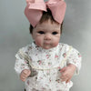A lifelike 18-inch reborn baby doll with hand-painted details wearing a white floral dress and pink bow