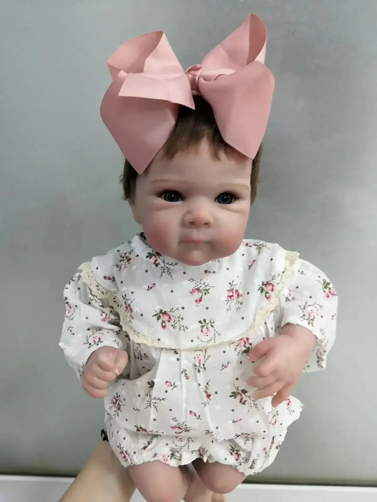 A lifelike 18-inch reborn baby doll with hand-painted details wearing a white floral dress and pink bow