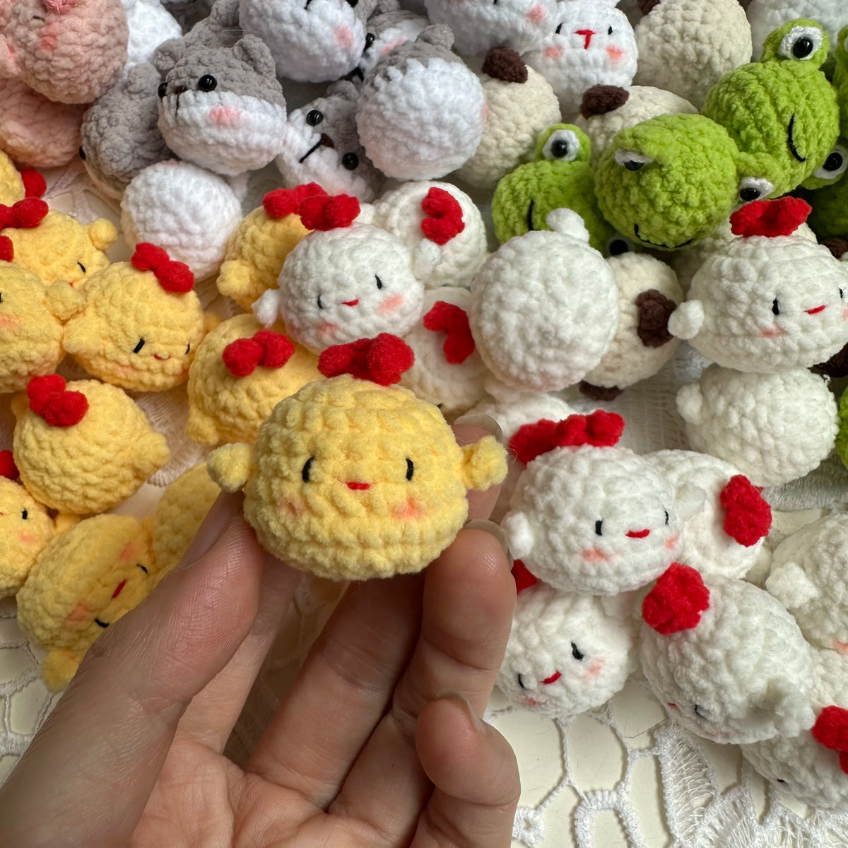 A hand holds a small handcrafted crochet animal keychain, likely a rabbit or chick, among many similar soft toys.
