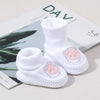 A pair of white knitted baby booties with pink embroidery and non-slip soles resting on a light surface.