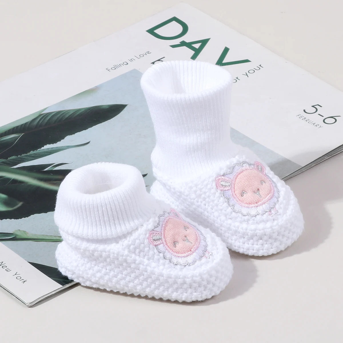 A pair of white knitted baby booties with pink embroidery and non-slip soles resting on a light surface.